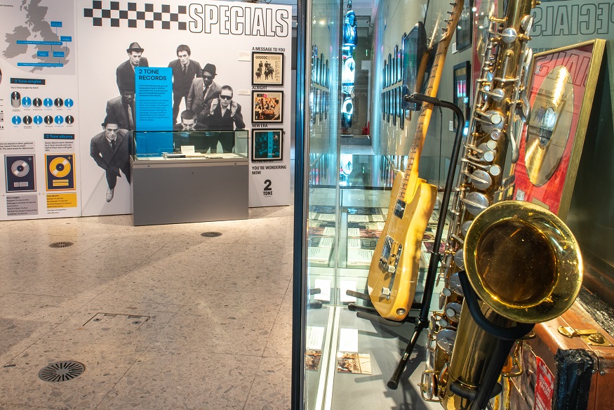 Image of an exhibition space – on the back wall are vinyl album covers and a large black and white photograph of a band wearing suits and sunglasses, below large text reading ‘The Specials’; on the left is a glass cabinet with yellow guitar, gold saxophone, brown suitcase and gold-disc framed record. 