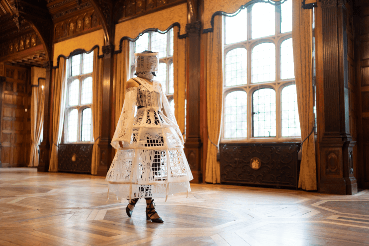 An image of a person wearing a dress, made up of an intricate paper design. The person is walking through a space with wooden floors and grand wooden beams, and large windows are behind them.