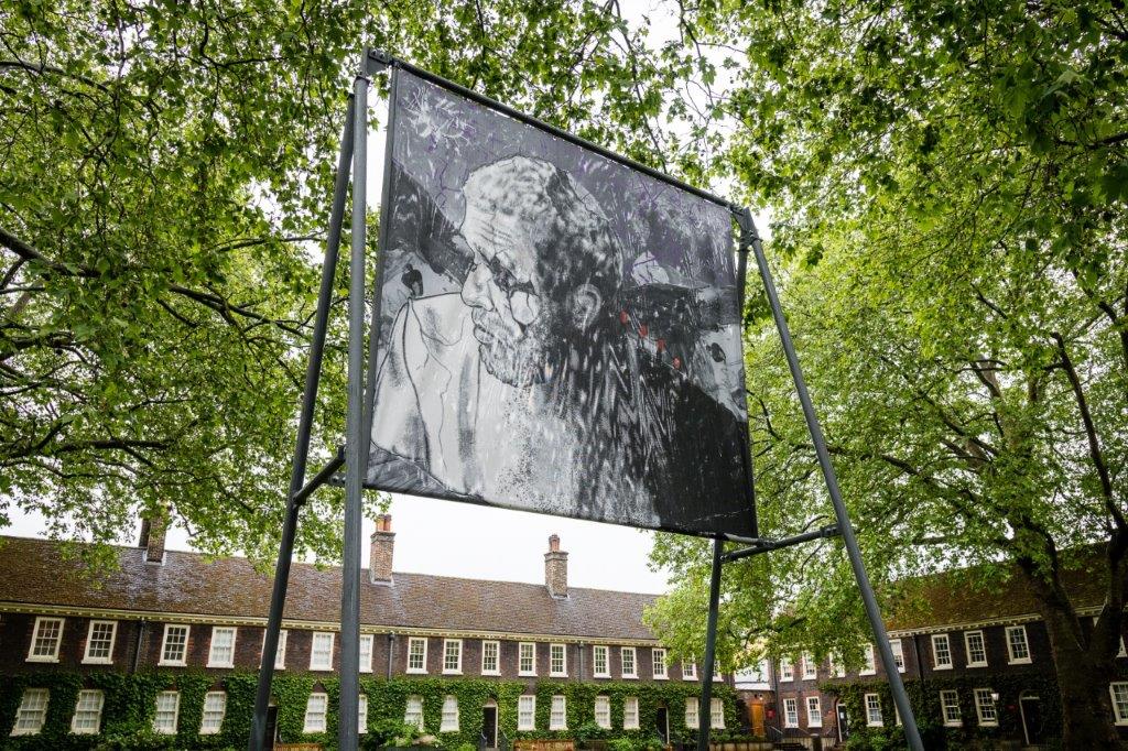 An image of a large billboard-like structure placed outside a large, ivy-covered building, and beneath the shelter of a few large trees. The structure shows a piece of artwork – a black and white illustration of a person.
