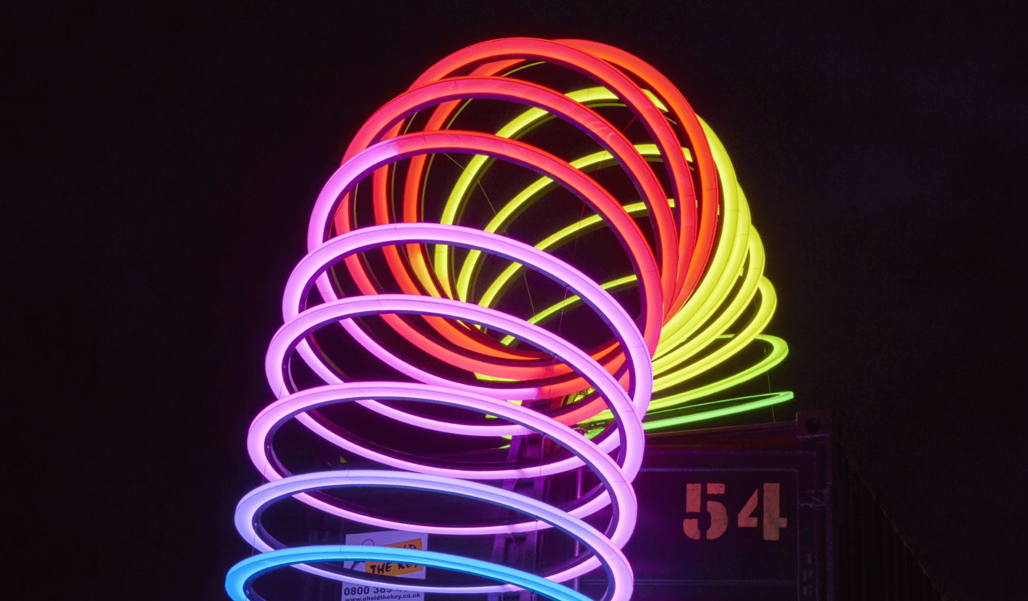 An illuminated sculpture of a large slinky-like spring is bended and sits on on top of a large, metal container – this piece feature at Durham Lumiere’s light.