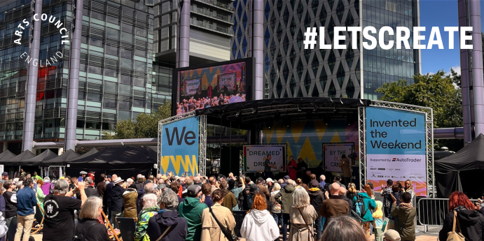 A crowd at a stage outside, the stage has a sign that says 'We Invented the Weekend'.