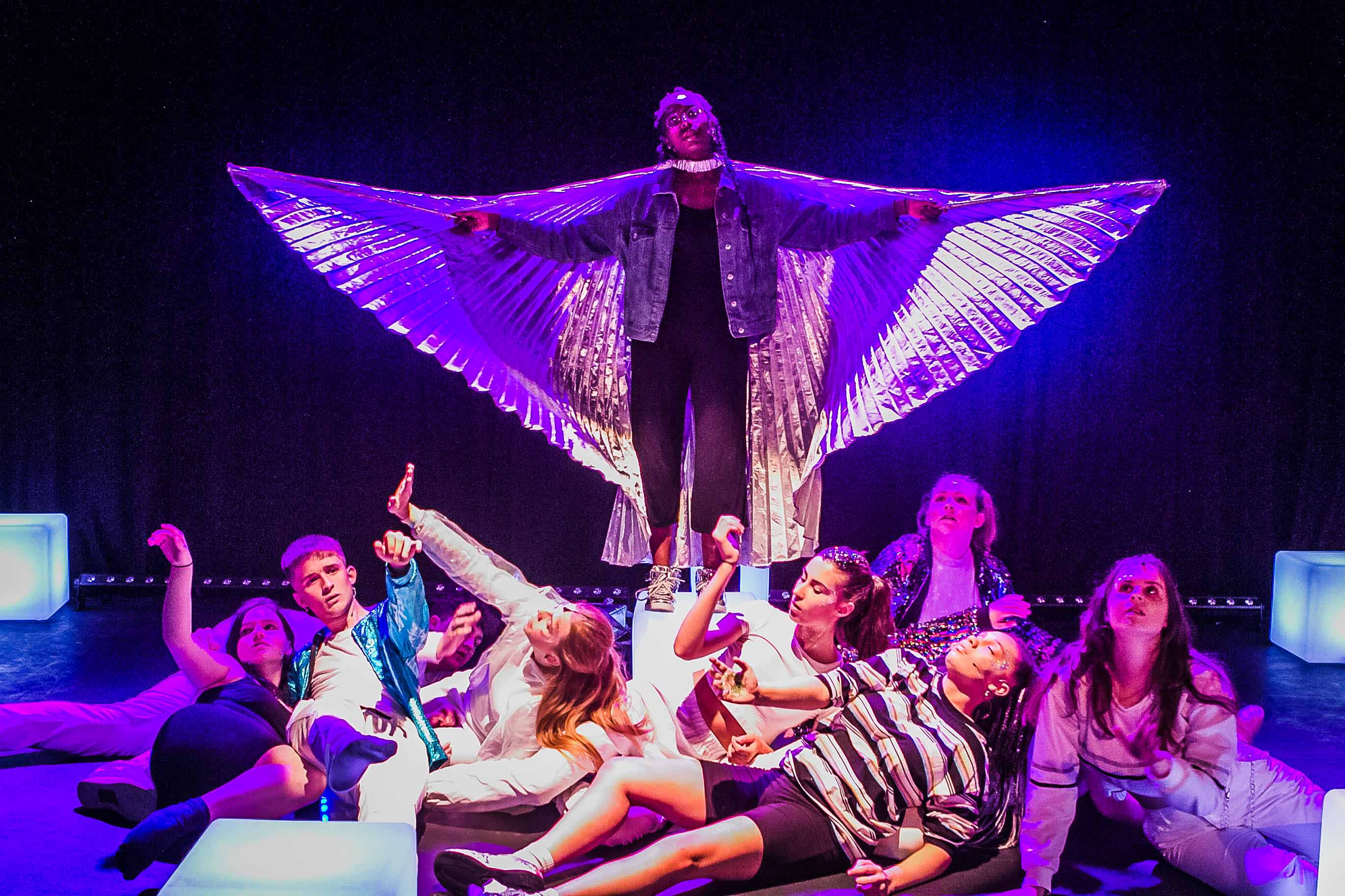 Young female, black actress (She/Her) wearing silver angel wings, surrounded by ensemble actors from the production.