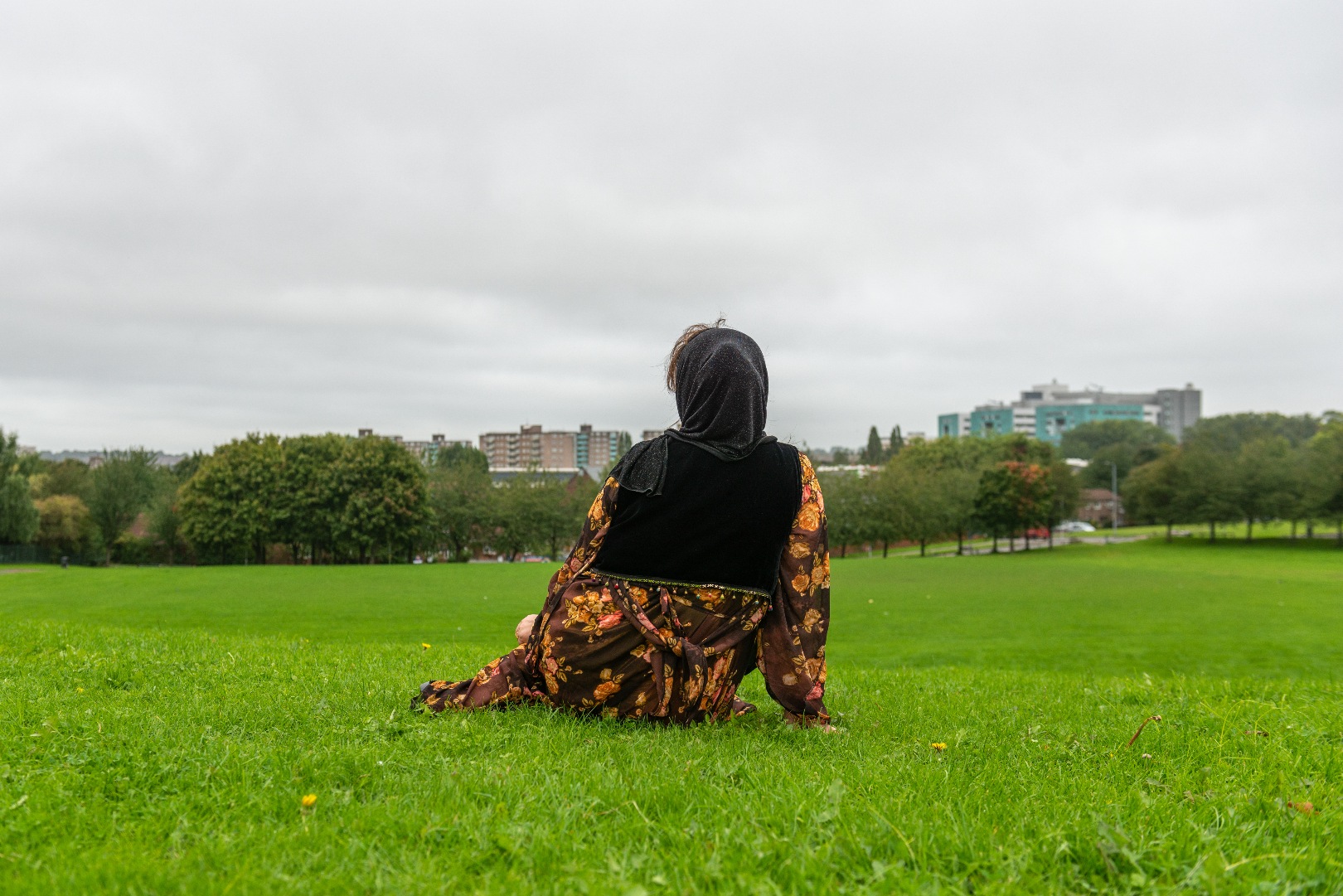 A woman wearing a floral robe and a black hijab sits in the park and looks out at the city skyline of Leeds. 