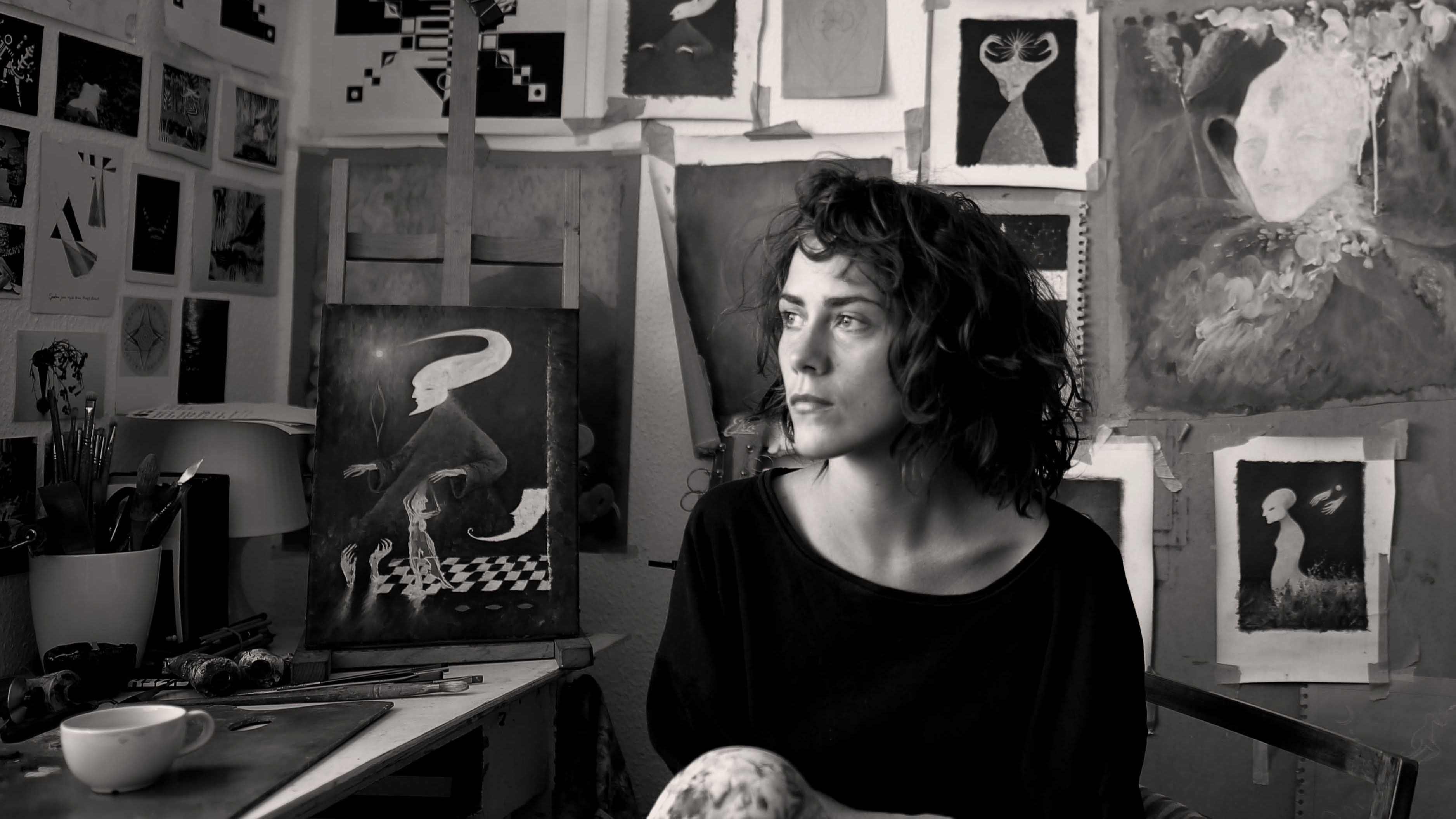 Black and white image of a woman with short dark curly hairy gazing out of a window. The wall behind her is covered in various paintings, drawings, and sketches. She is sat at a desk.
