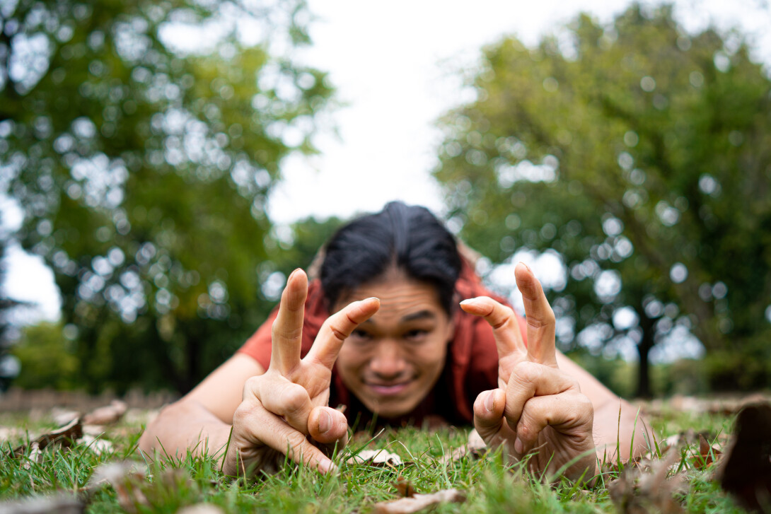 A photograph of a person crouching in the grass outdoors, making insect shapes with their fingers. 