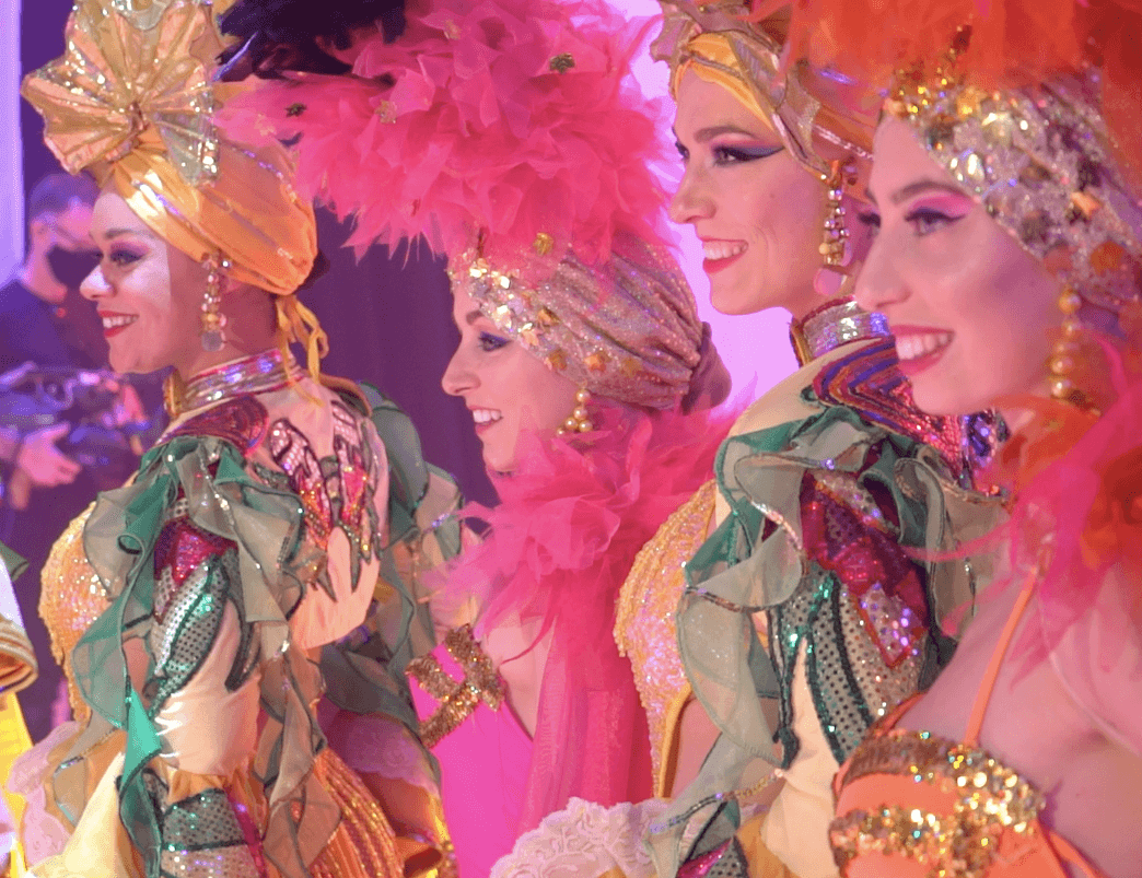 A photograph of four people. They wear ornate outfits with gold sequins and frills, and pink feathered head wear.