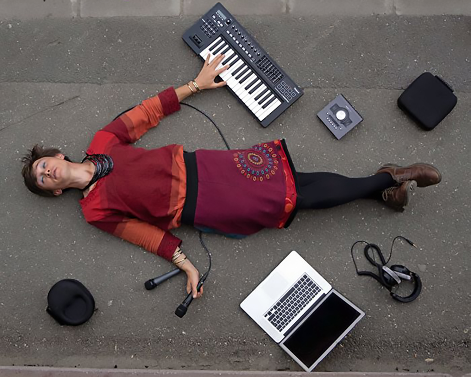 Photograph of a person laying down. Next to them is a keyboard, a laptop, and a microphone.