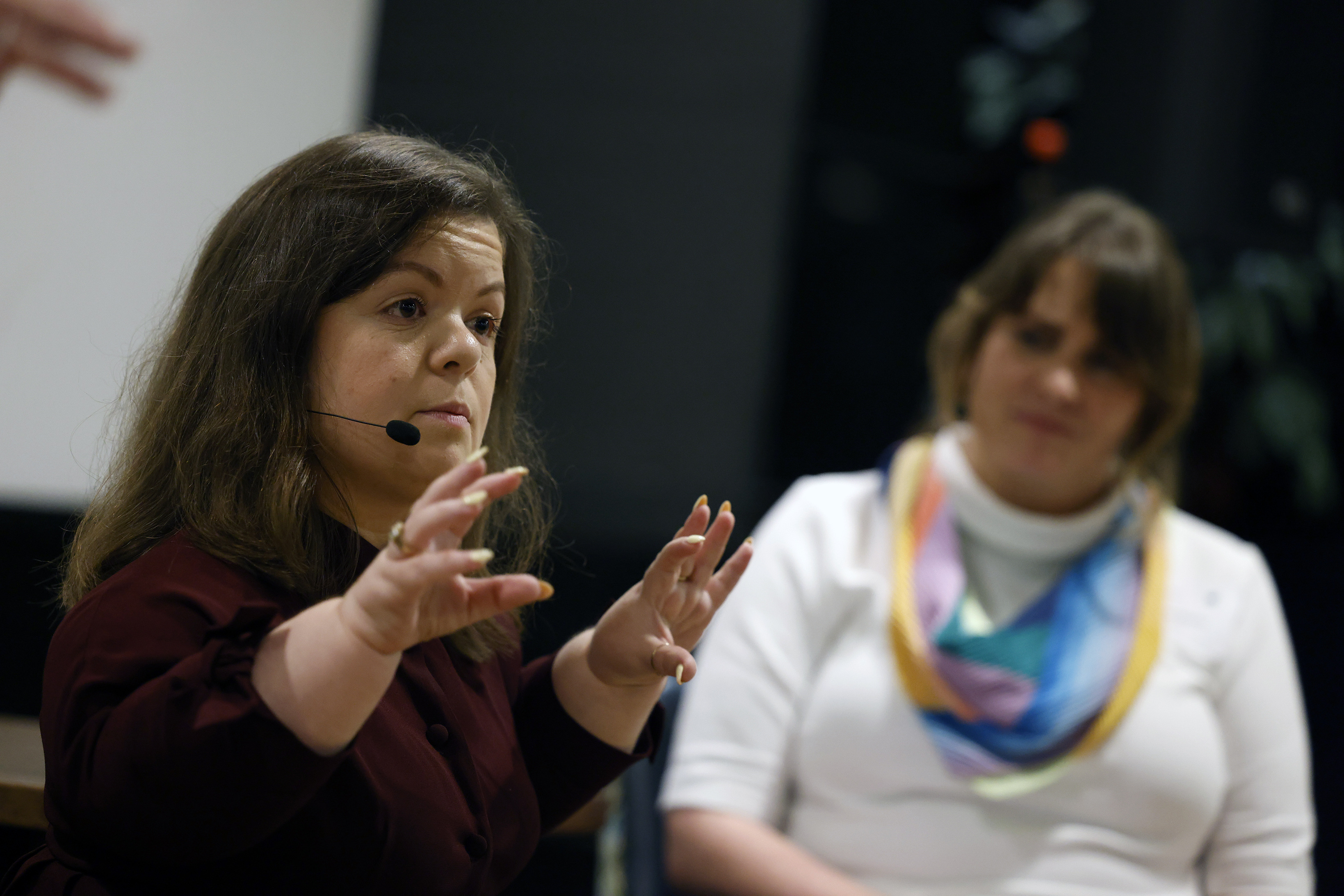 A woman speaking into a headset microphone gestures with both hands while giving a talk; another woman in a colourful scarf sits blurred in the background.