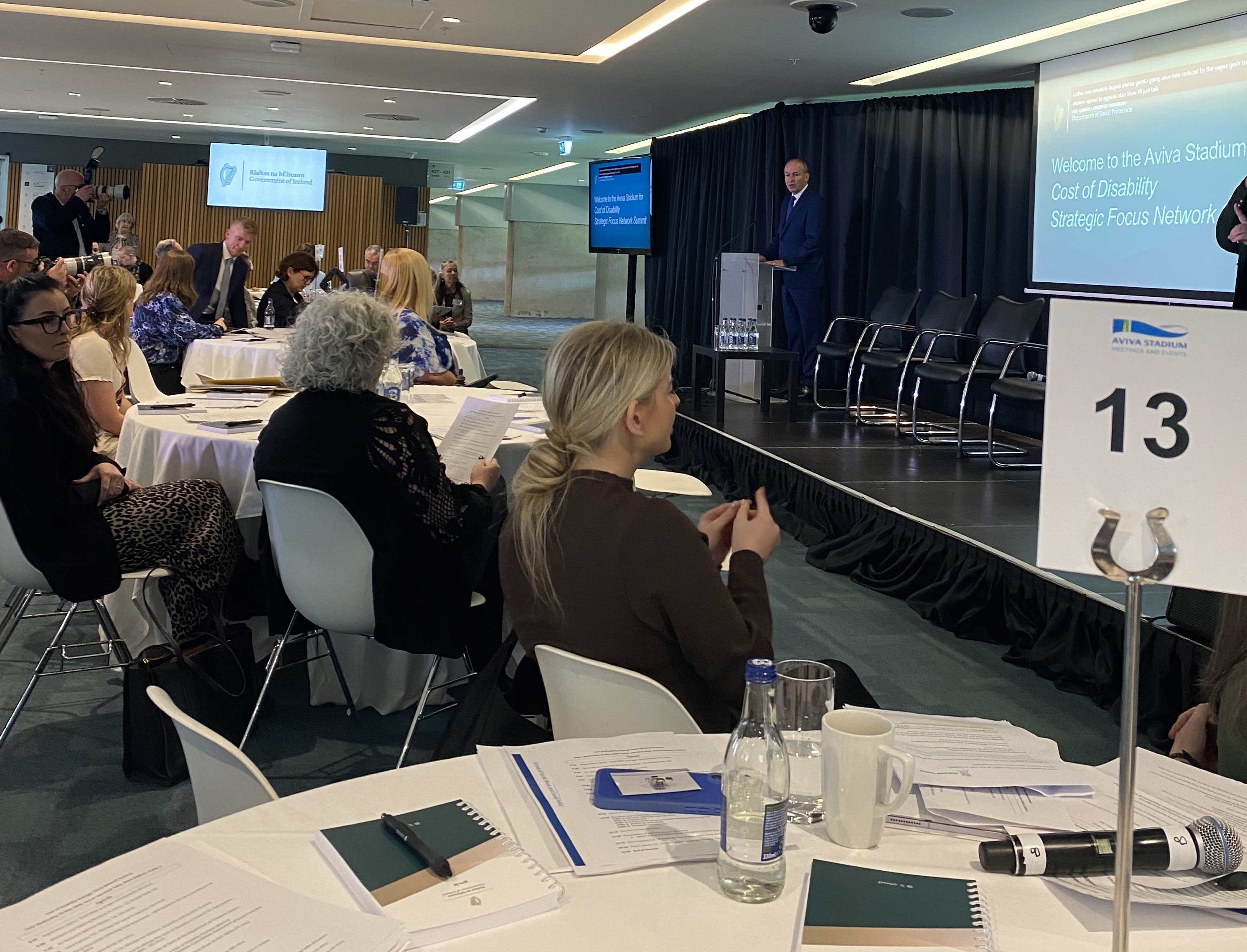 An Taoiseach addresses delegates at the Cost of Disability Strategic Focus Network Summit at Aviva Stadium. Attendees are seated at numbered round tables with documents and materials in front of them. A Government of Ireland logo is visible on screen at the frontA speaker addresses delegates at the Cost of Disability Strategic Focus Network Summit at Aviva Stadium. Attendees are seated at numbered round tables with documents and materials in front of them. A Government of Ireland logo is visible on screen at the front