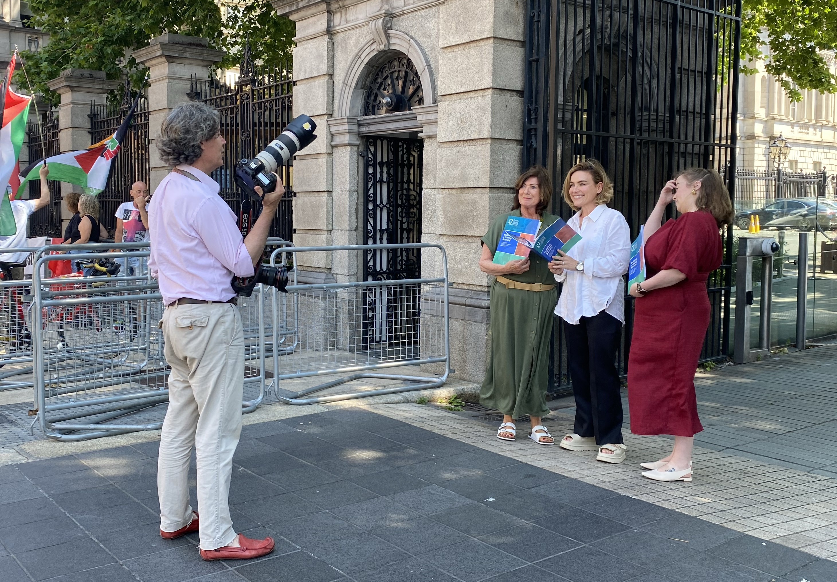On a city street, a photographer with a large telephoto lens films three women standing by railings outside a building. One woman holds a document. A Palestinian flag is visible in the background.