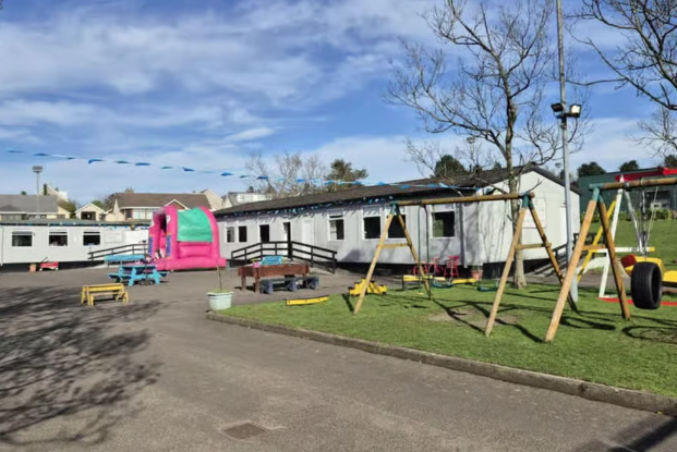 An outdoor play area at a single-storey prefab-style building. A pink bouncy castle, wooden swings, a tyre swing, picnic benches and bunting are set out on grass and tarmac under a blue sky.
