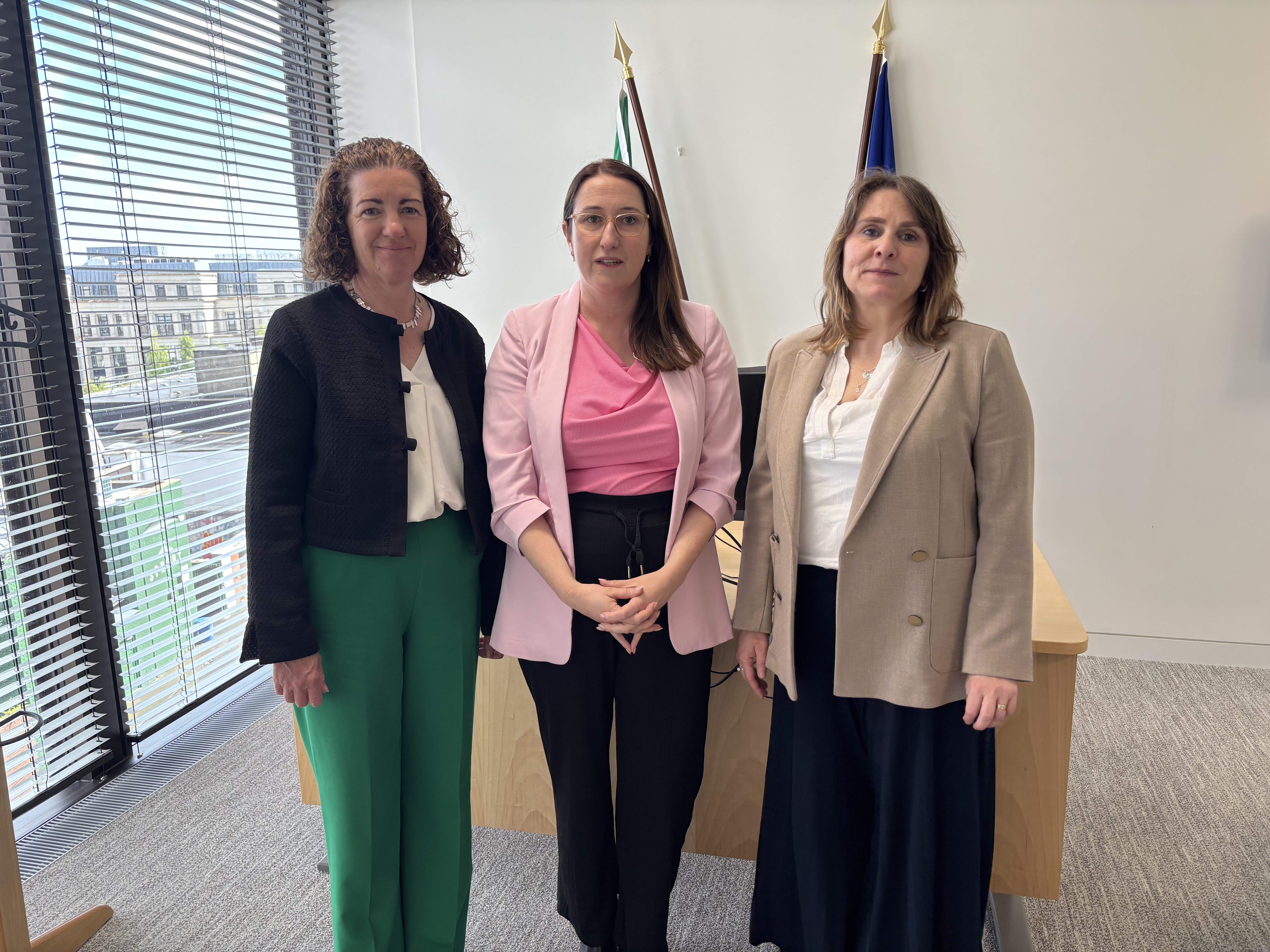 Three women standing together in a bright office setting with Irish and EU flags visible in the background.
