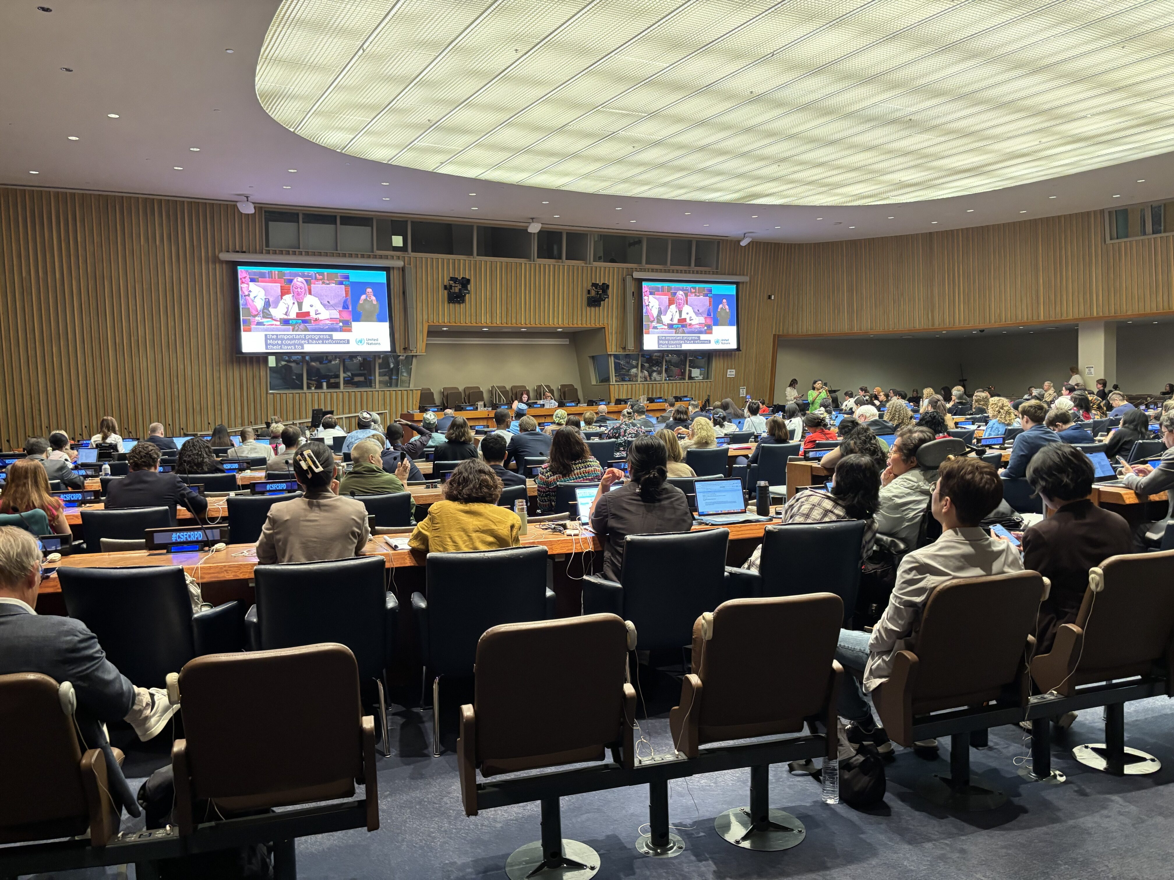 Wide view of a UN conference hall filled with delegates, with two large screens showing a speaker and an Irish Sign Language interpreter.