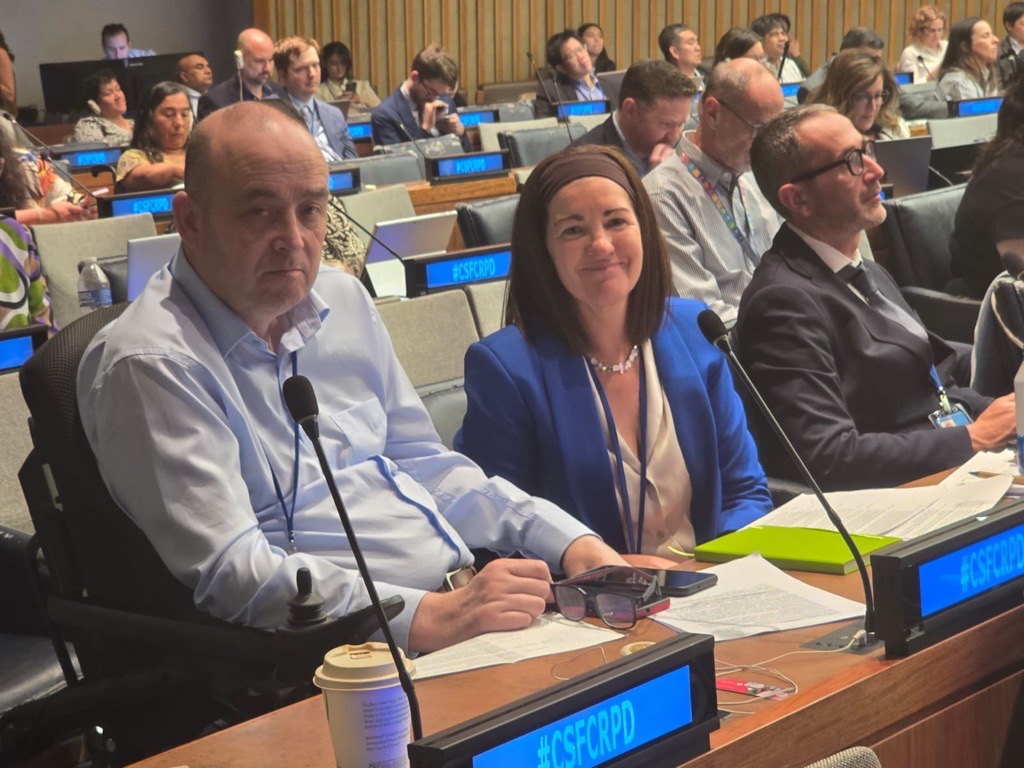 A man in a wheelchair and two colleagues seated at a UN conference desk with 