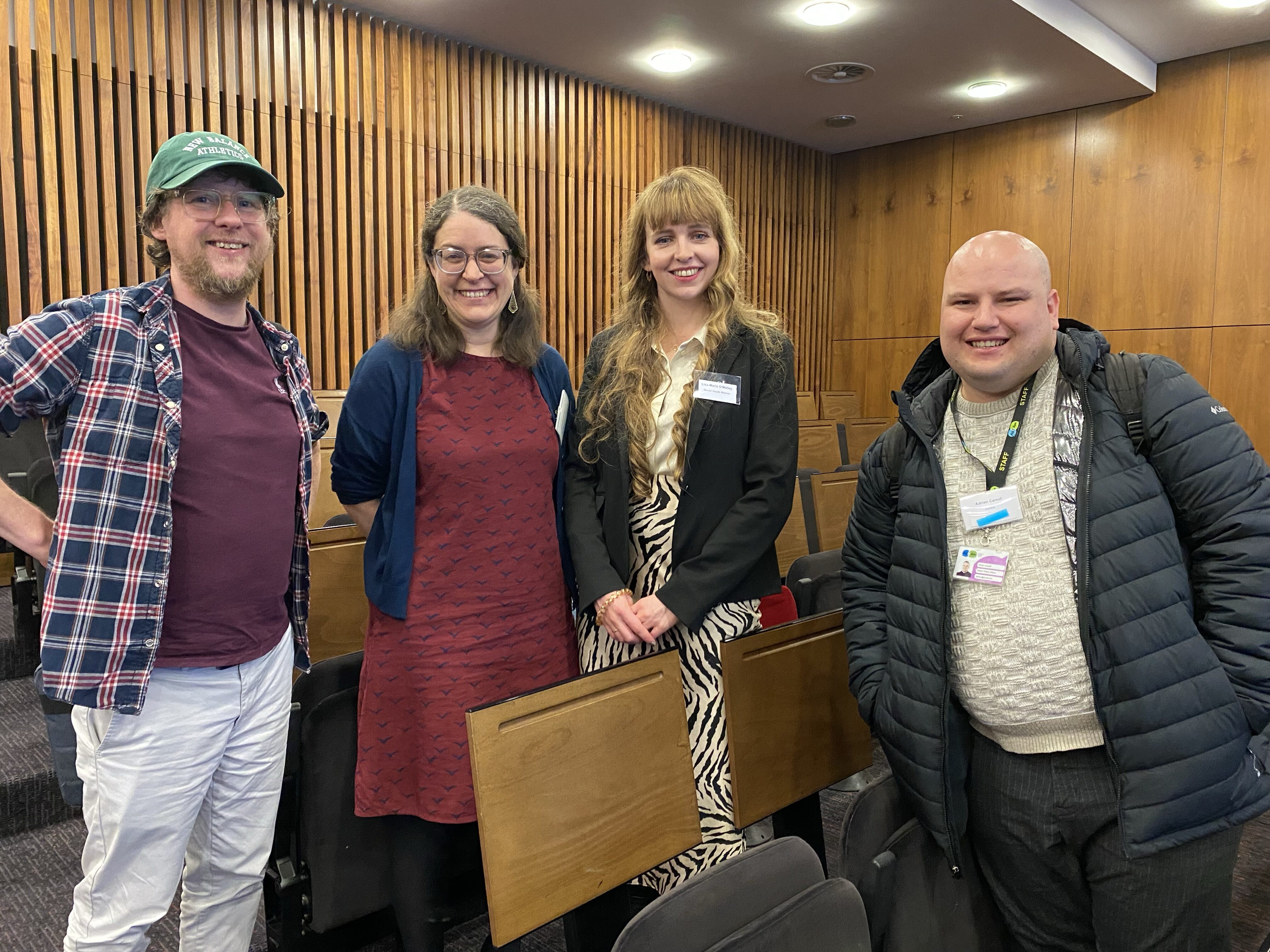Four people stand smiling together in a wood-panelled lecture theatre. From left: a man in a green cap and red plaid shirt, a woman with glasses in a navy cardigan and red dress, a woman with long blonde hair in a black blazer and zebra-print outfit, and a man in a dark puffer jacket wearing a lanyard.