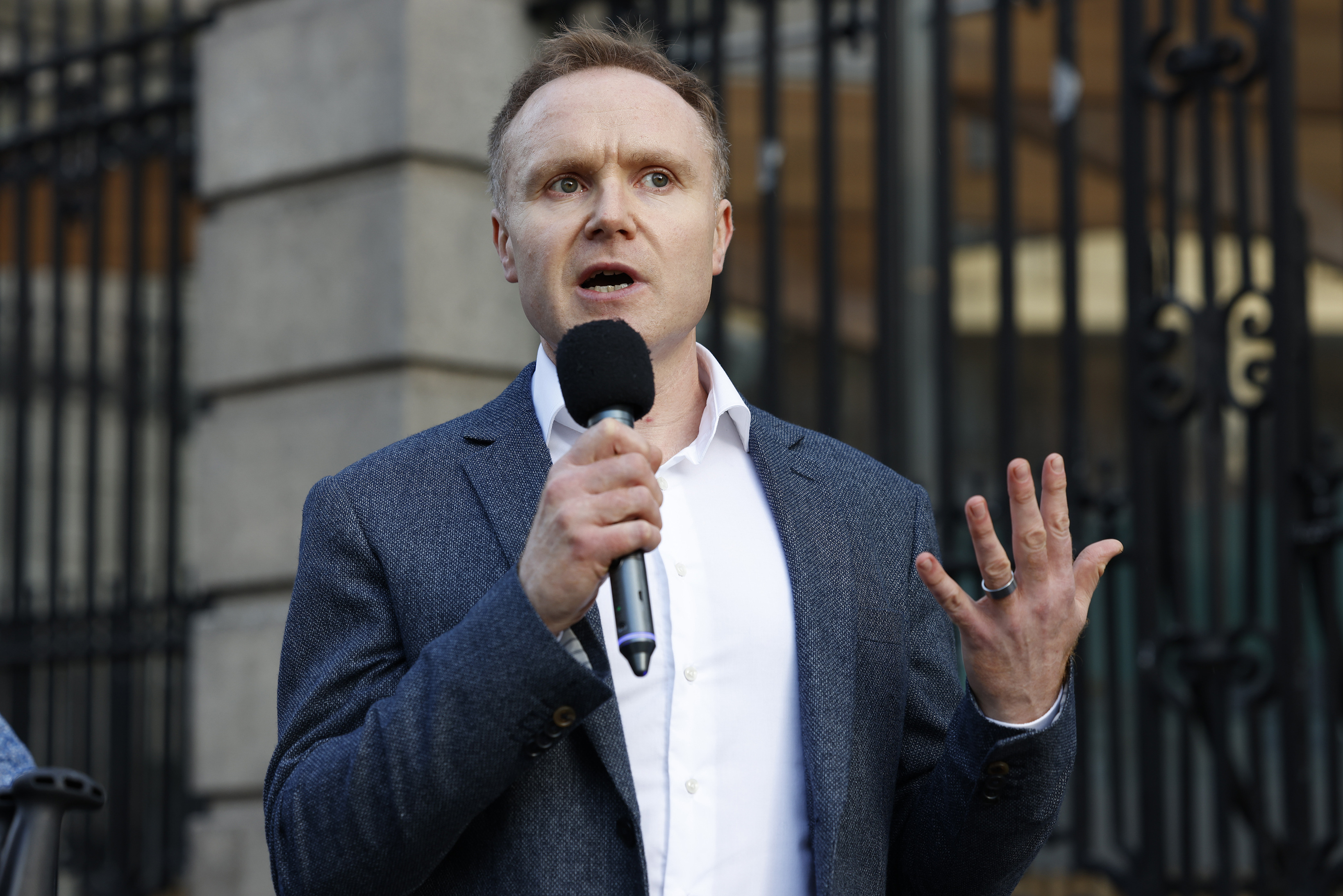 A man in a blazer speaking into a microphone and gesturing, standing outside a building with iron railings.