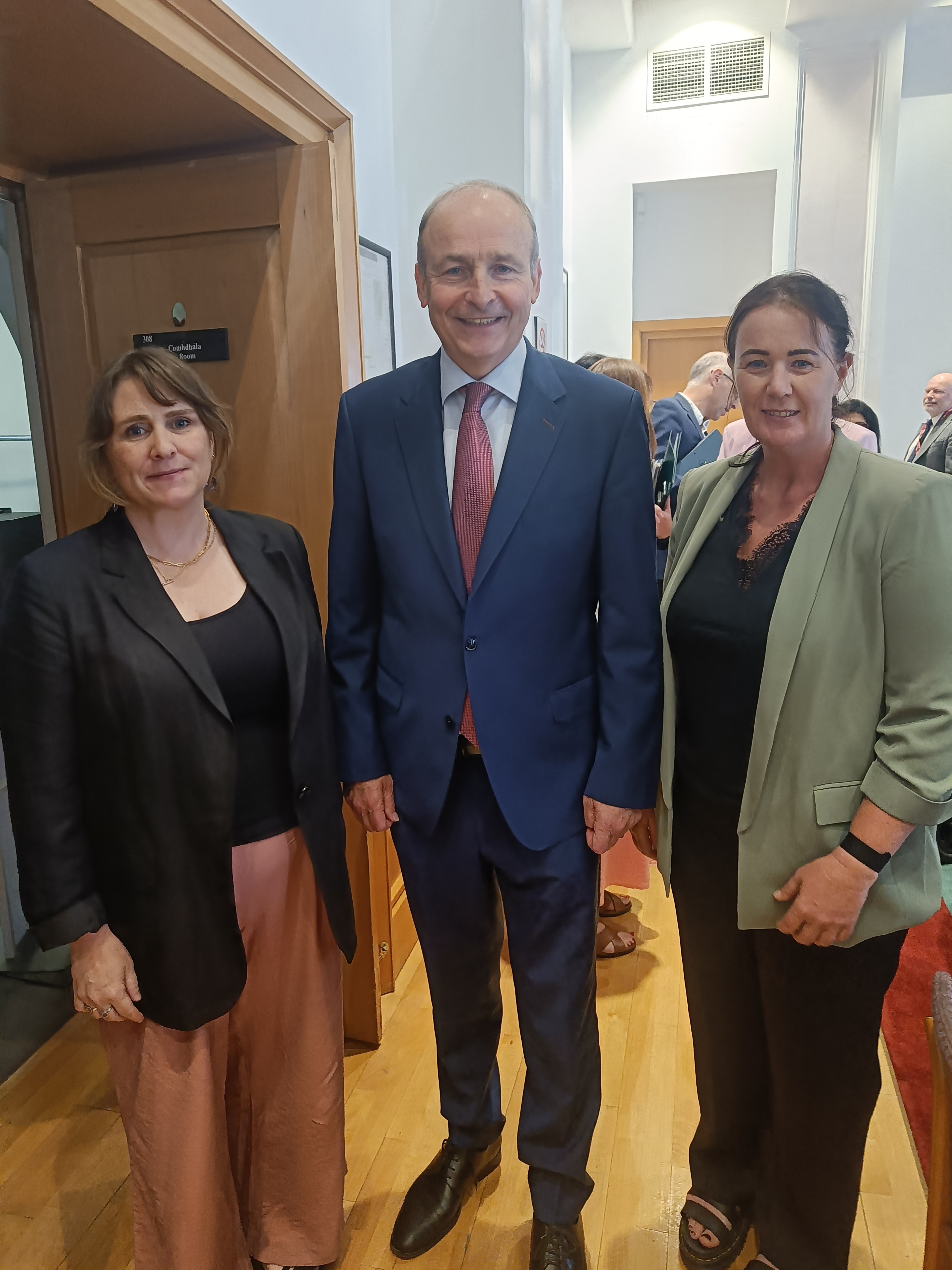 Three people pose for a photo indoors. An Taoiseach Micheal Martin is in a dark suit and red tie stands in the centre between DFI Director of Advocacy Emer Begley and Karen Foley, Chair of the NDSA, Emer is in a black blazer and pink trousers, Karen is in a green blazer and black top.