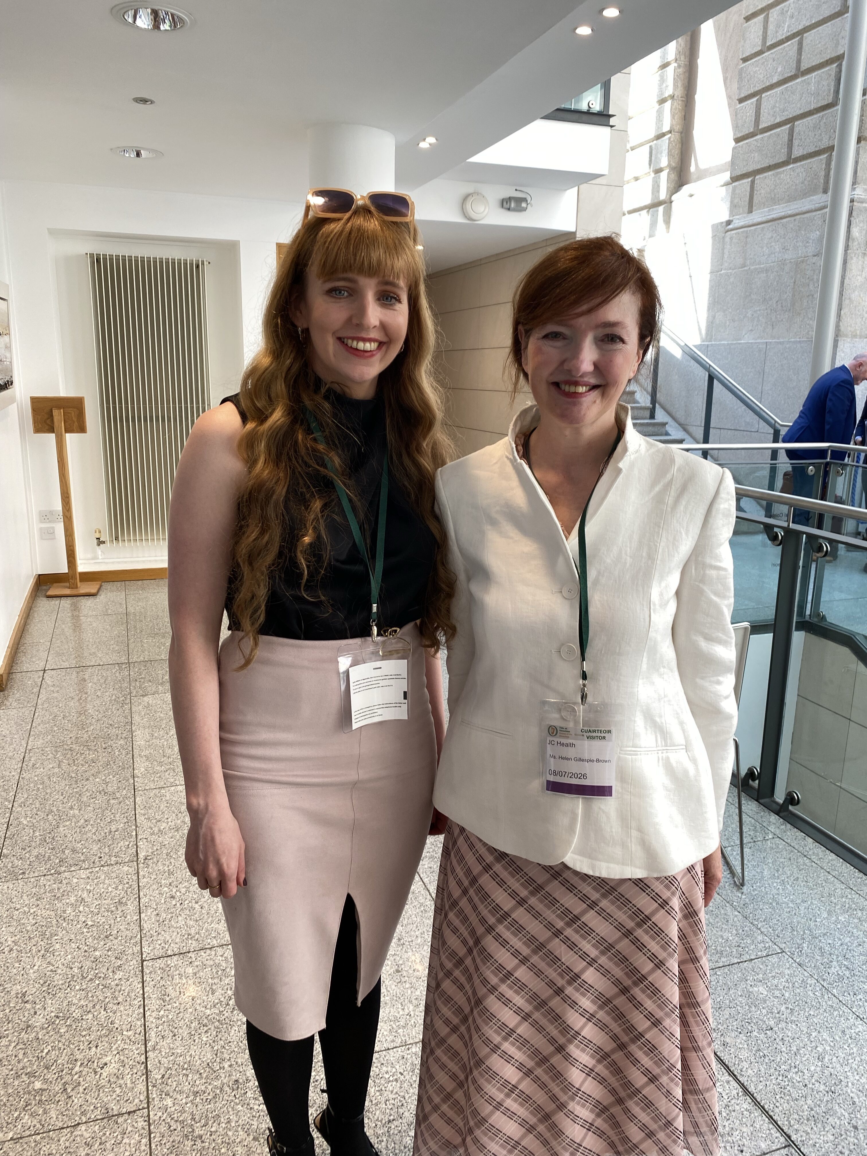 Two women stand smiling together in a bright stone-floored building. One has long wavy hair and wears a black top with a pink skirt; the other wears a white blazer and tartan skirt. Both wear lanyards.