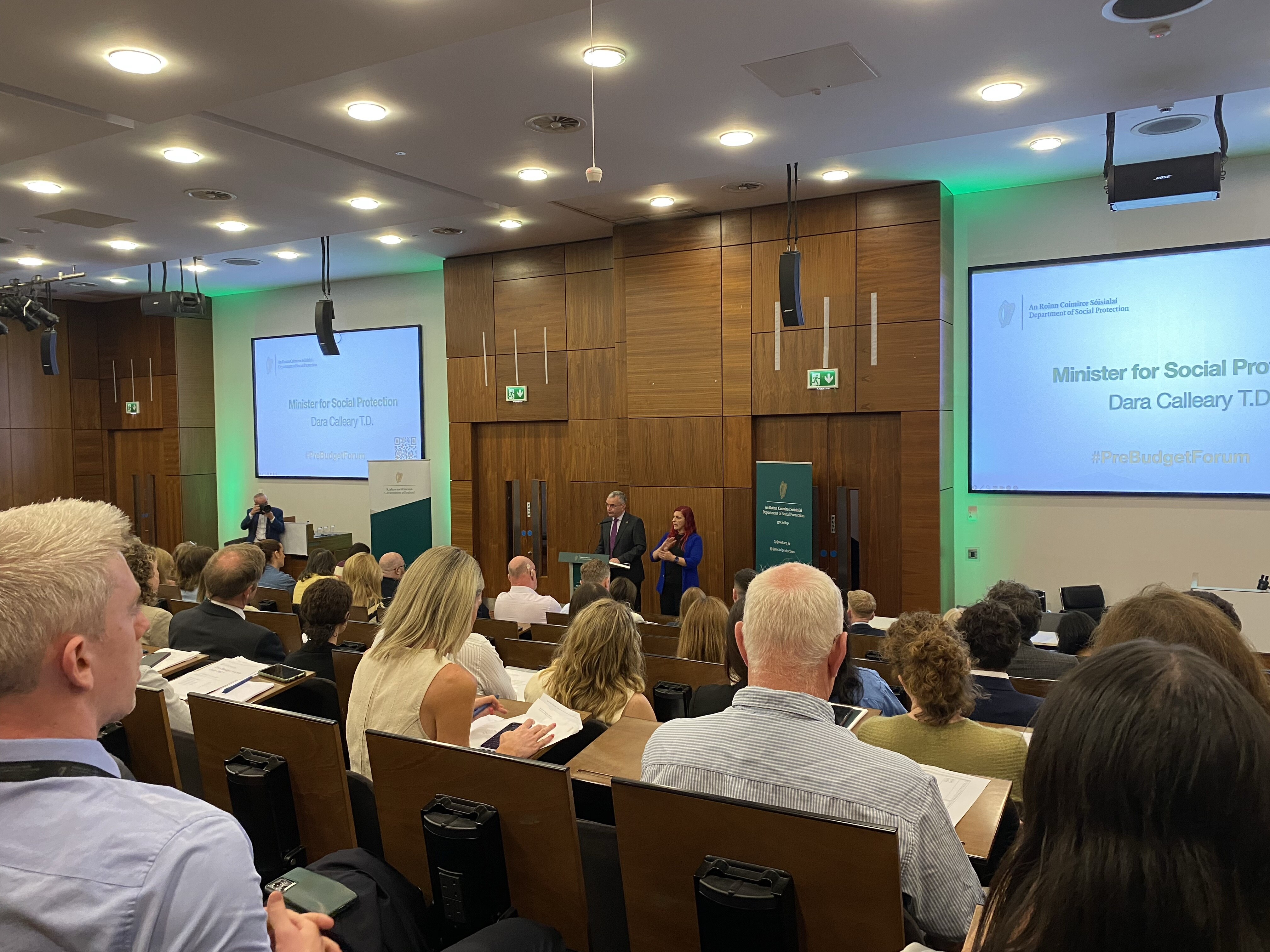 A conference or seminar in a modern wood-panelled lecture theatre. A speaker stands at a lectern beside a woman, with a seated audience facing them. A presentation screen reads 