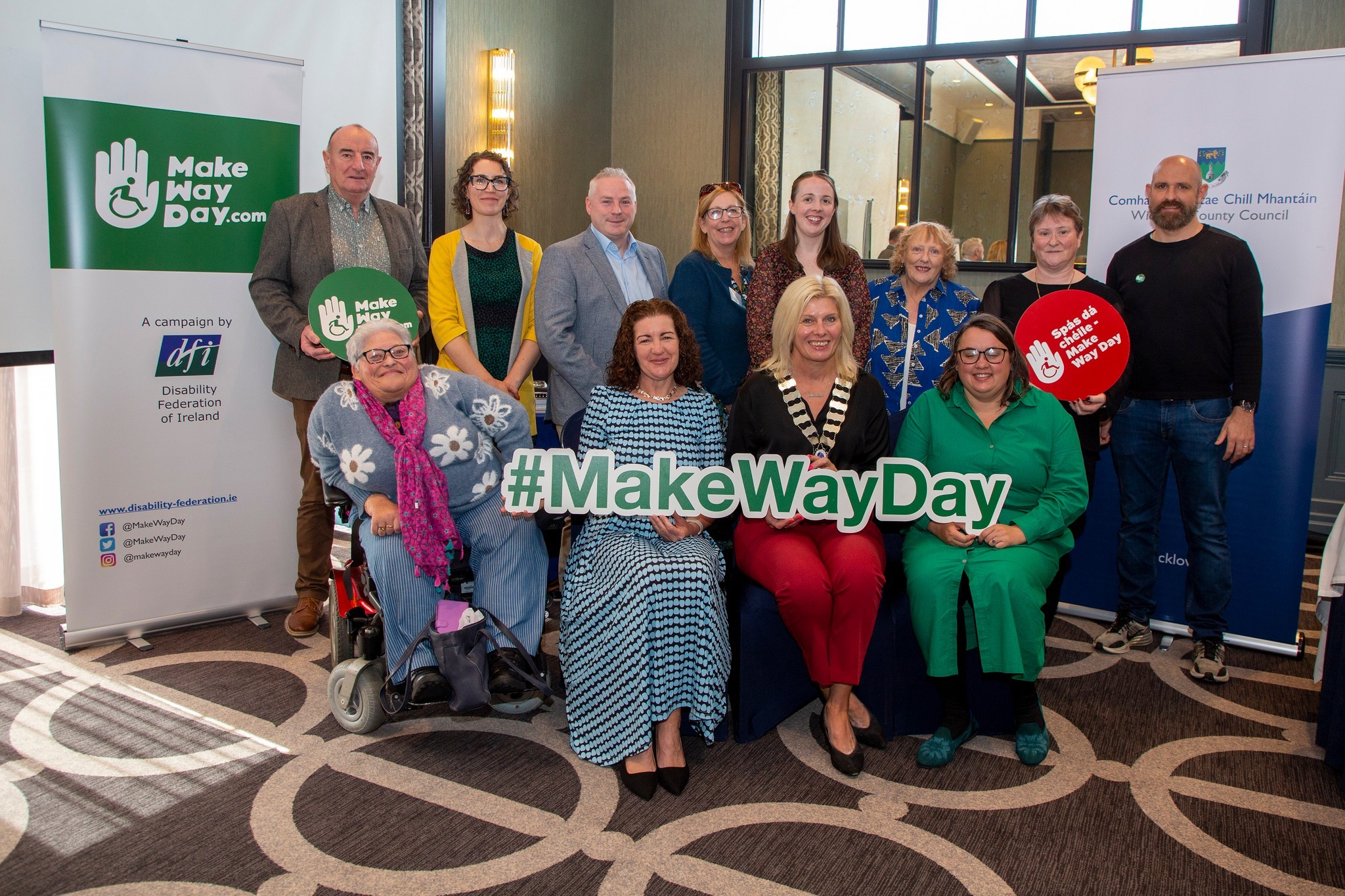 10:21 A group of thirteen people pose together at the Make Way Day launch event in Carlow, with a DFI Make Way Day banner on the left and a Carlow County Council banner on the right. A #MakeWayDay hashtag sign is displayed in front of the group, which includes people standing and seated, one of whom is a wheelchair user.