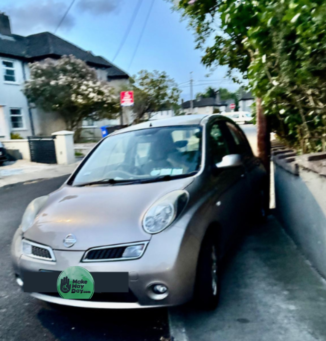 A gold/beige Nissan Micra parked on a residential street beside a hedge, with houses in the background. A green 