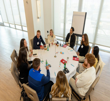 An overhead view of a group of people seated around a round table at a workshop or networking event, with notebooks, drinks and materials on the table, beside large windows.