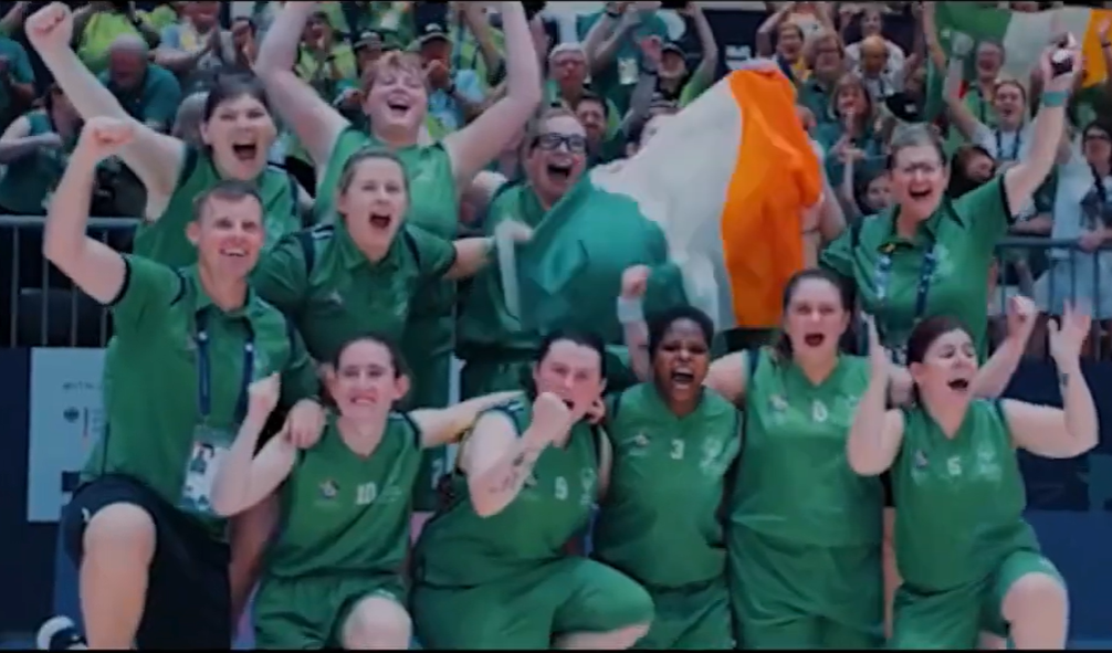 A cheering team in green Ireland jerseys celebrating together, with a tricolour flag and crowd in the background.